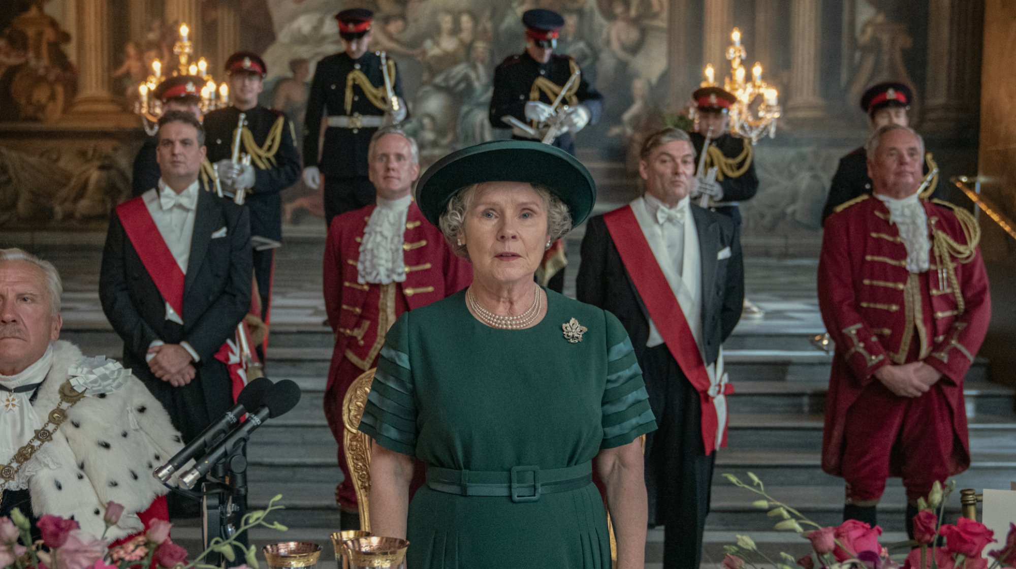 An actor resembling Queen Elizabeth II stands with attendants at a royal occasion.