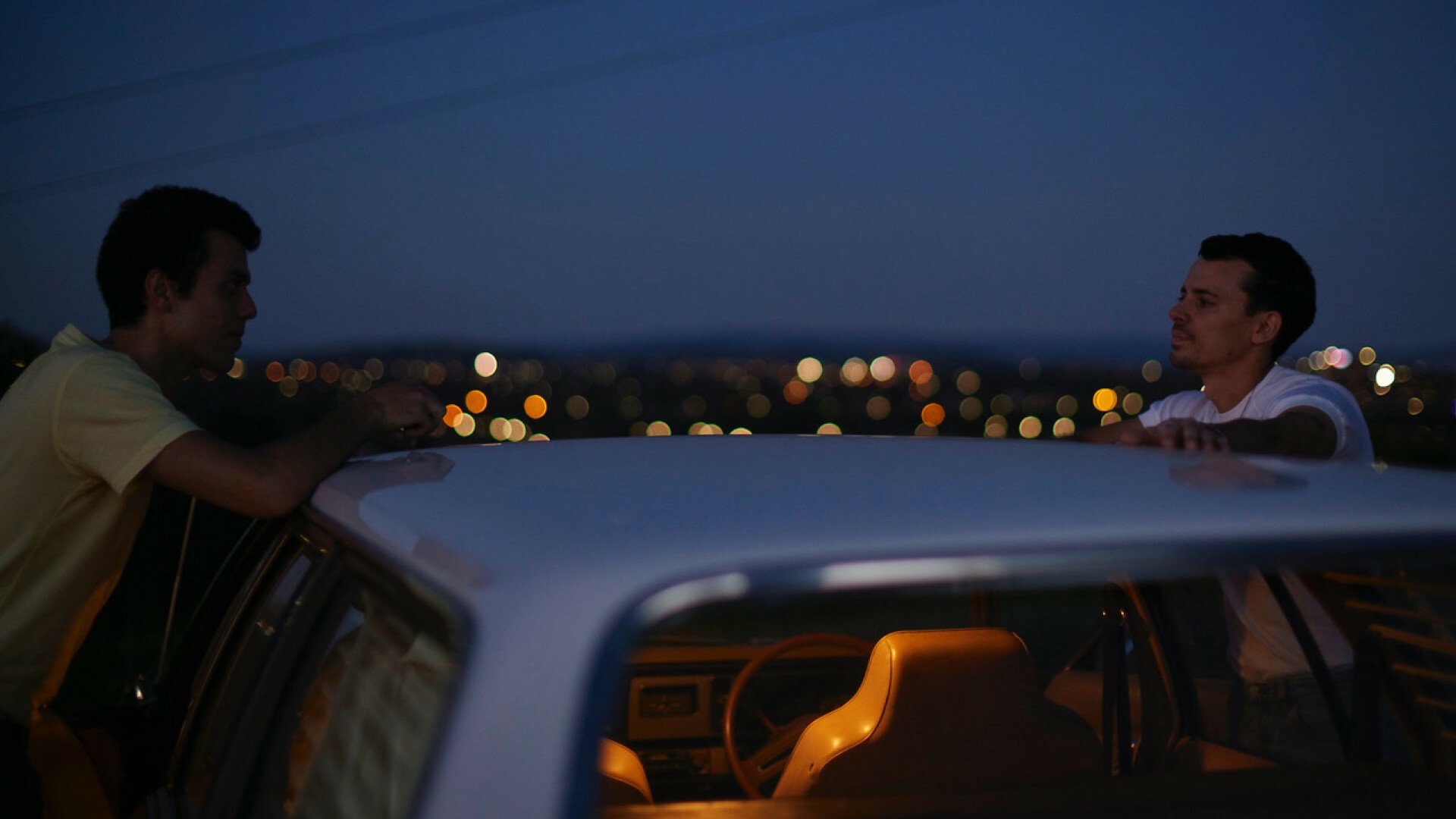 Two young men stand on either side of a car at night, looking at each other.