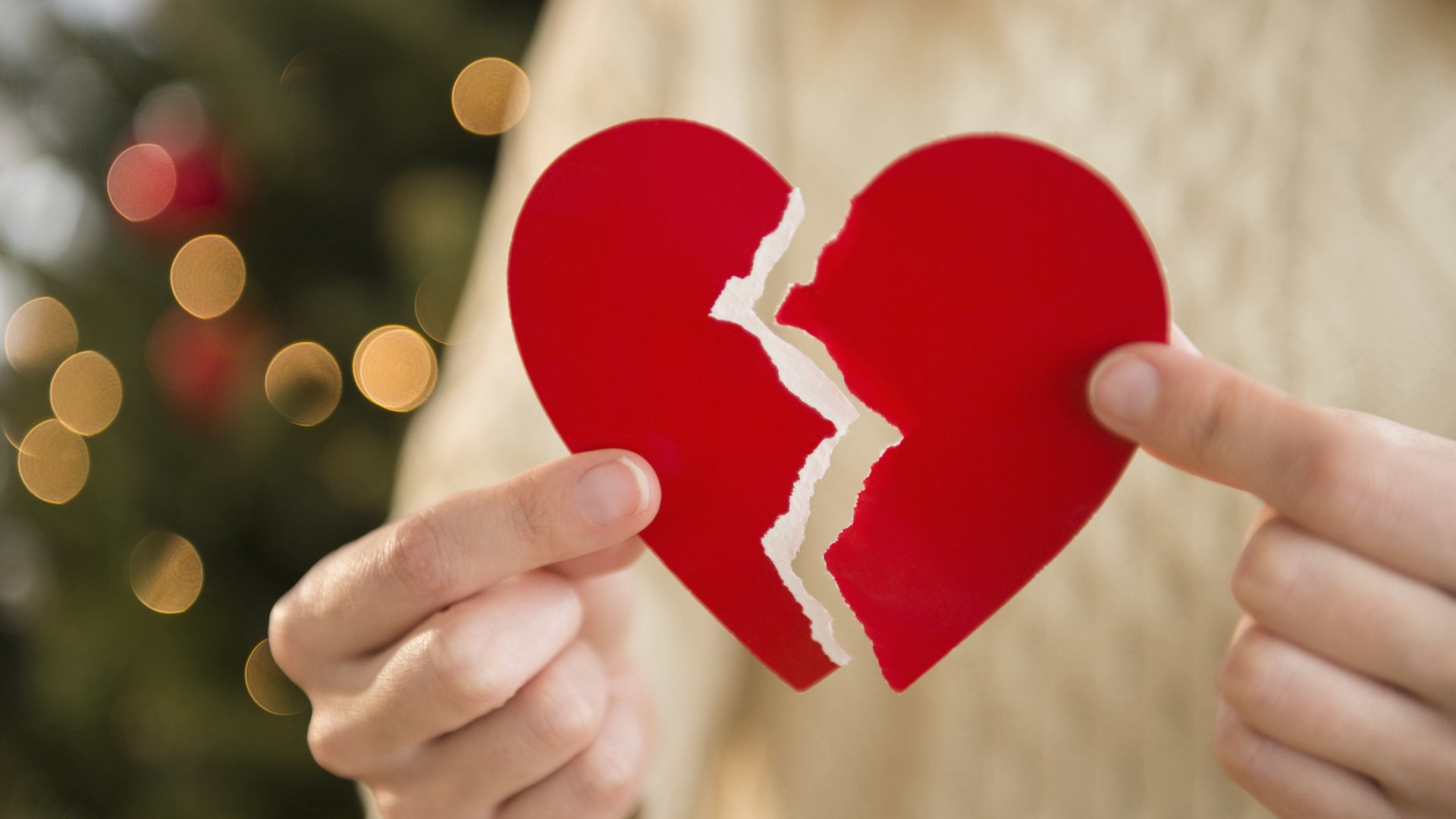 A person holds a paper heart with a rip down the middle, with a Christmas tree in the background.