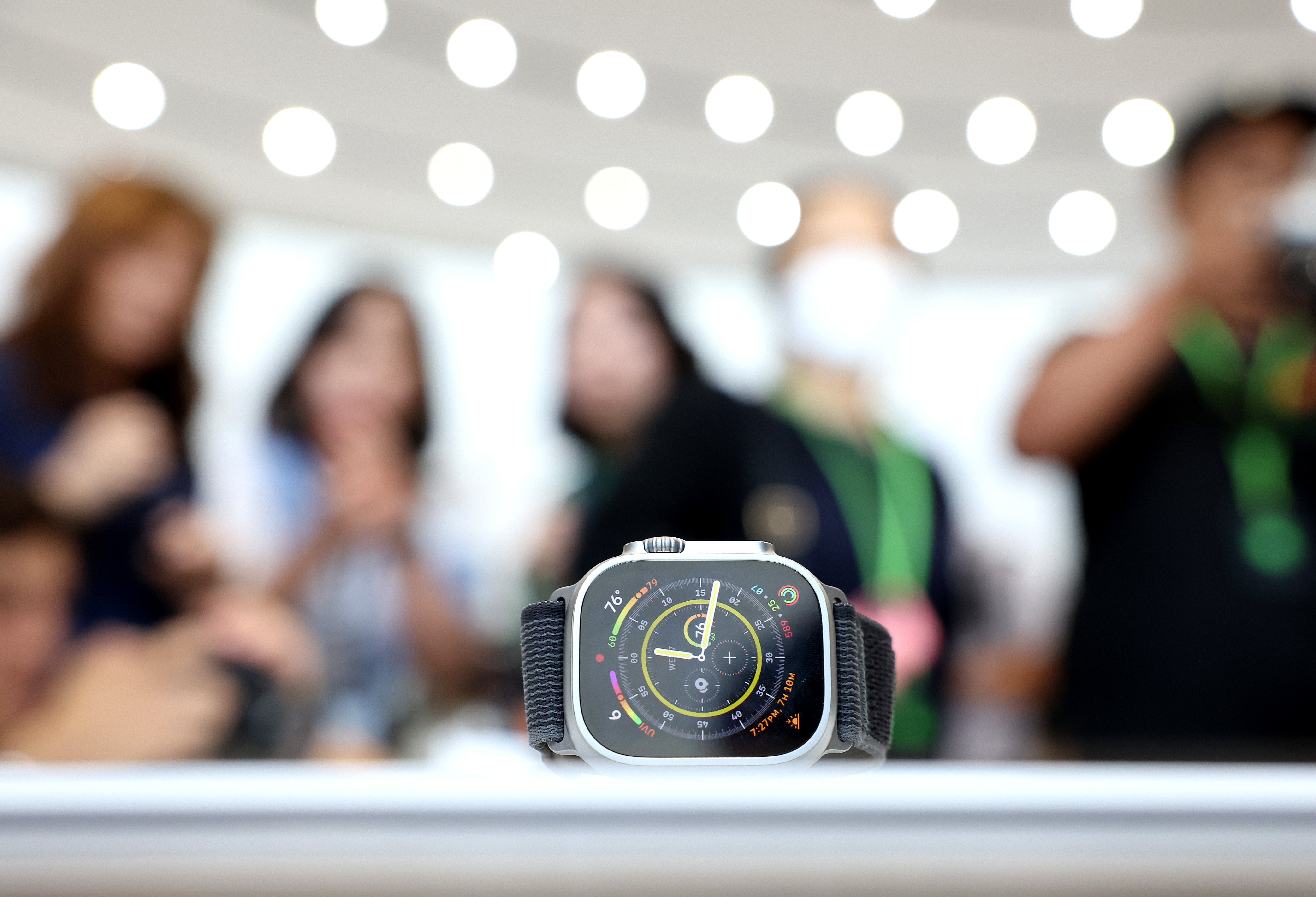 An apple watch rests on a white table in the middle of an Apple store. A group of customers are blurred out in the background.