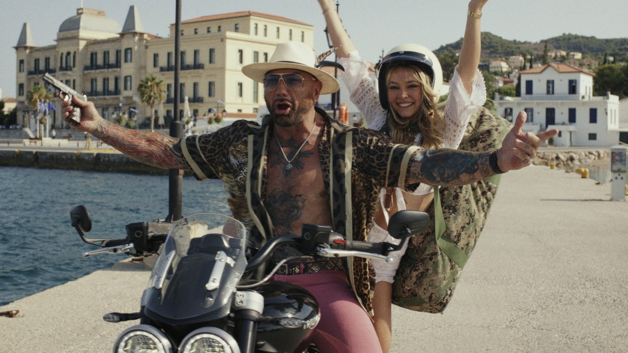 A man and a woman on a motorcycle ride up a dock. The man is holding a pistol.