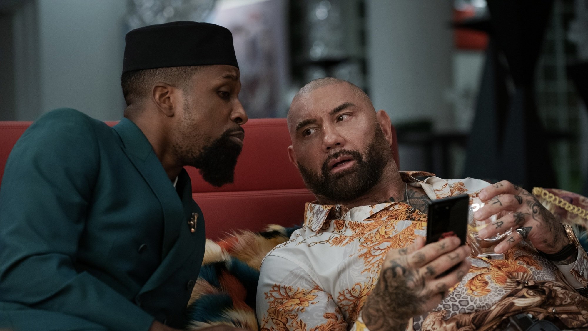 Two men sit on a red couch, looking at a phone.