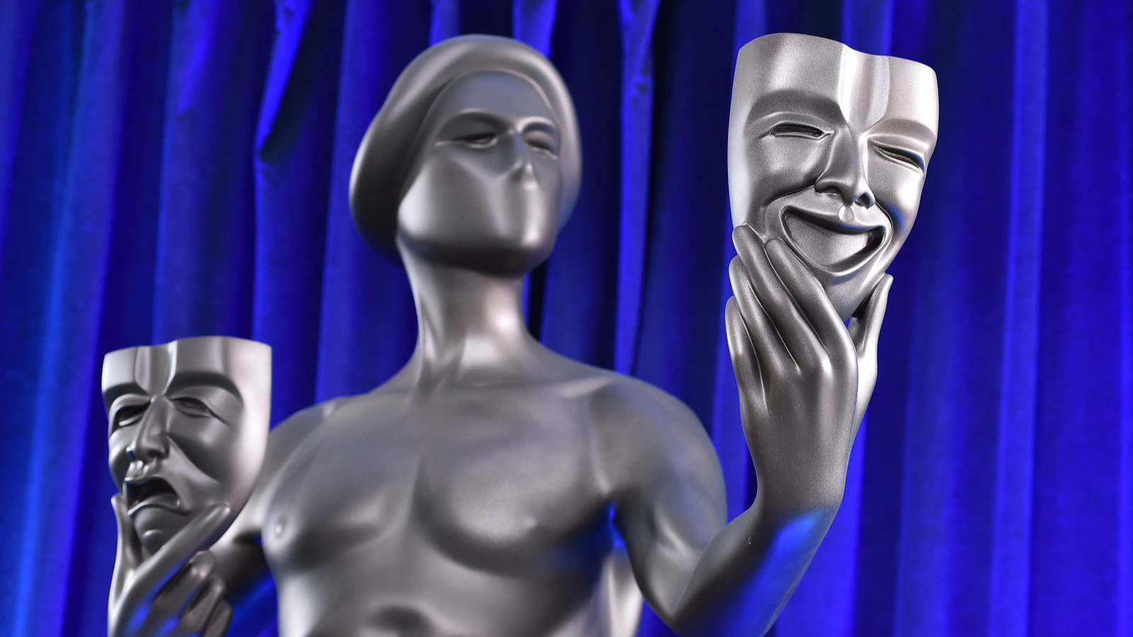 A metallic statue of a figure holding up two theater masks. 