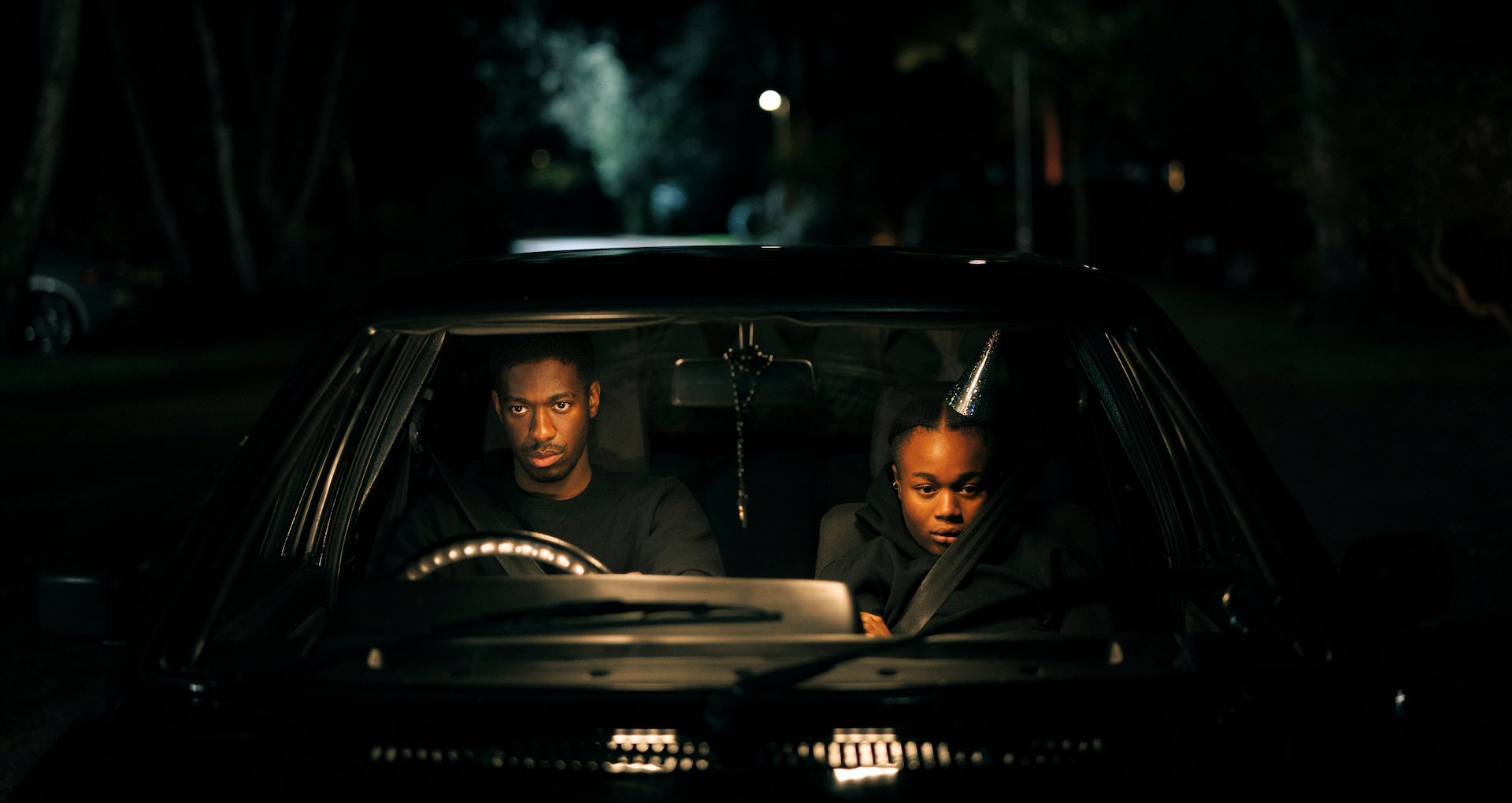 Two people sit in a parked car at night, one wearing a party hat, both looking serious.