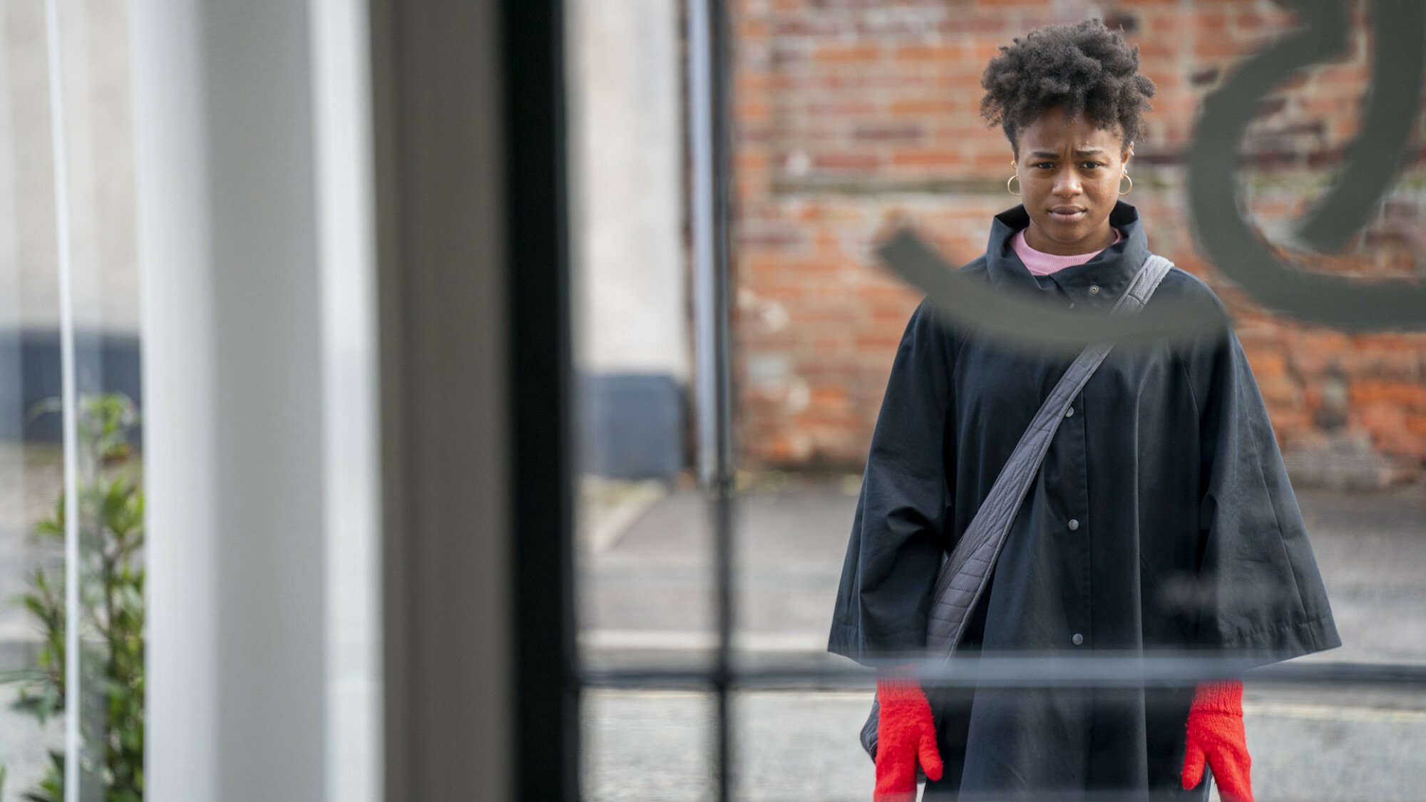 A young woman stands outside, seen through a window. She's facing the camera and wearing bright red gloves.