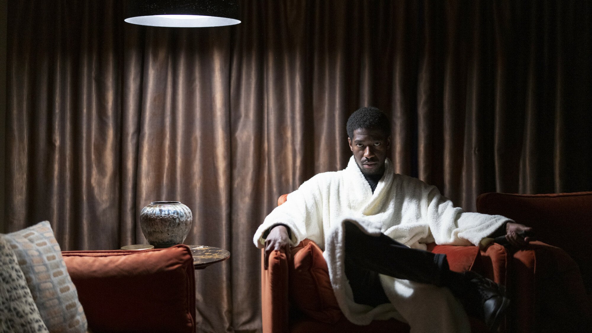 A man in a white robe sits in a stylish living room lit only by an overhead lamp.