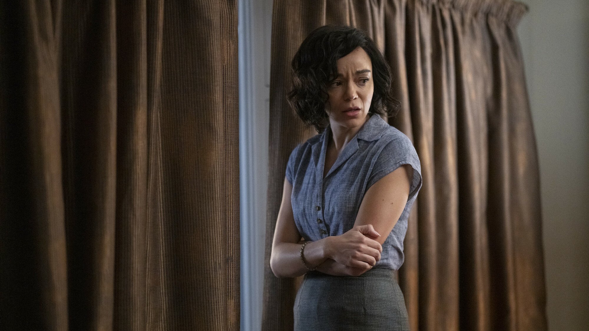 A woman looks nervous standing near a window with the curtains drawn.