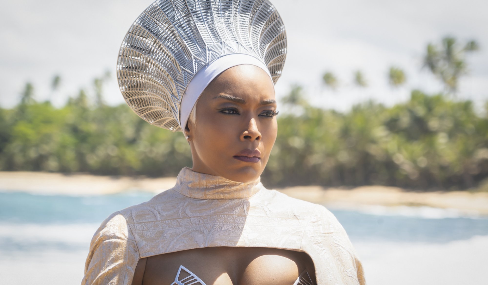 A woman in a silver crown and formalwear stands on an island beach looking serious.