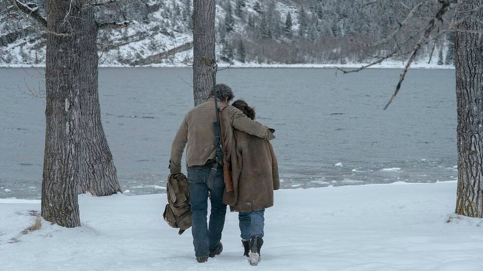 A man with his arm around a young girl walks away from the camera through the snow, towards a lake.
