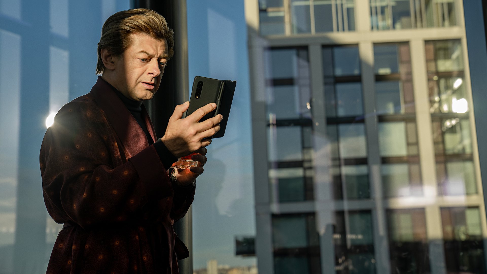 A wealthy man in a robe uses a foldable phone in an all glass apartment.