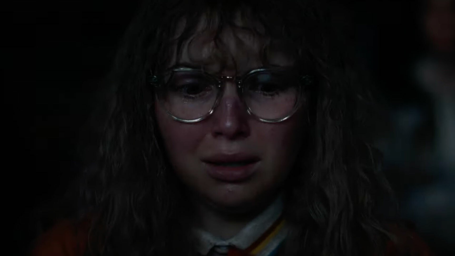 A woman with glasses stands in a dark room looking frightened.