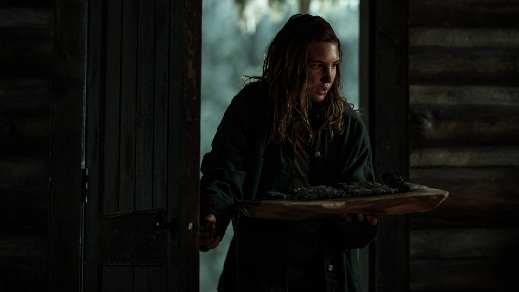 A teenage girl wearing a coat enters a cabin while holding a tray of meat. 