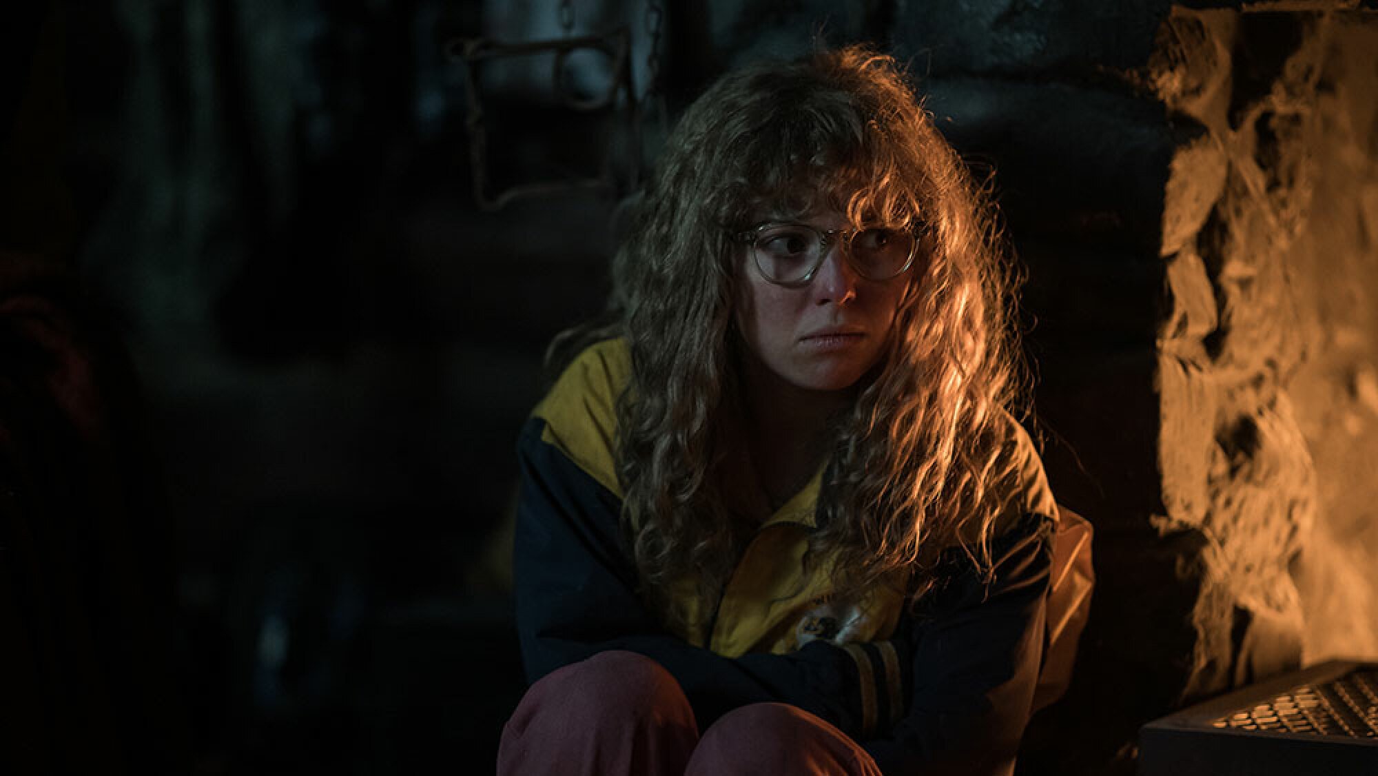 A girl with glasses and frizzy blonde hair sits by a fireplace.