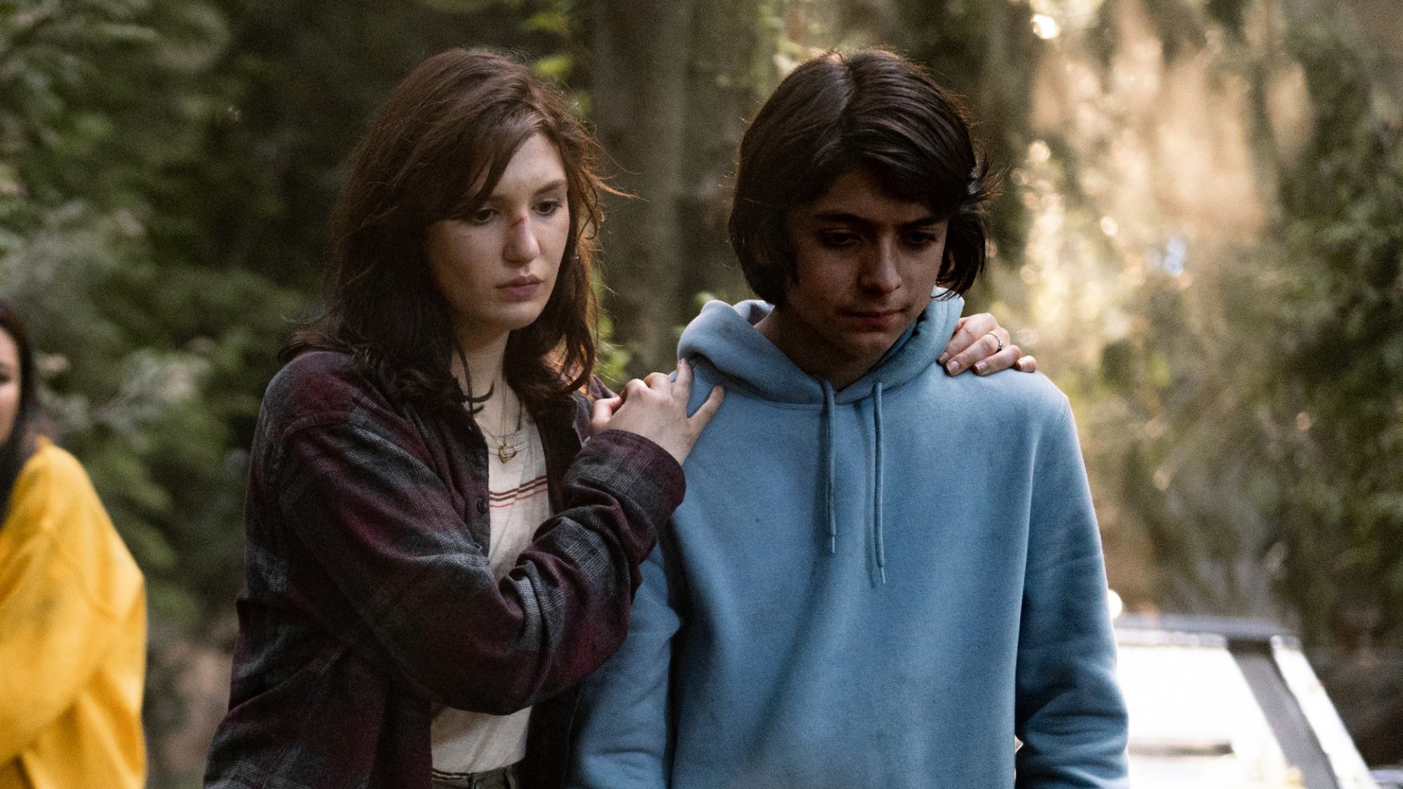 A teen girl holds a teen boy by the shoulders in the woods, both look solemn.