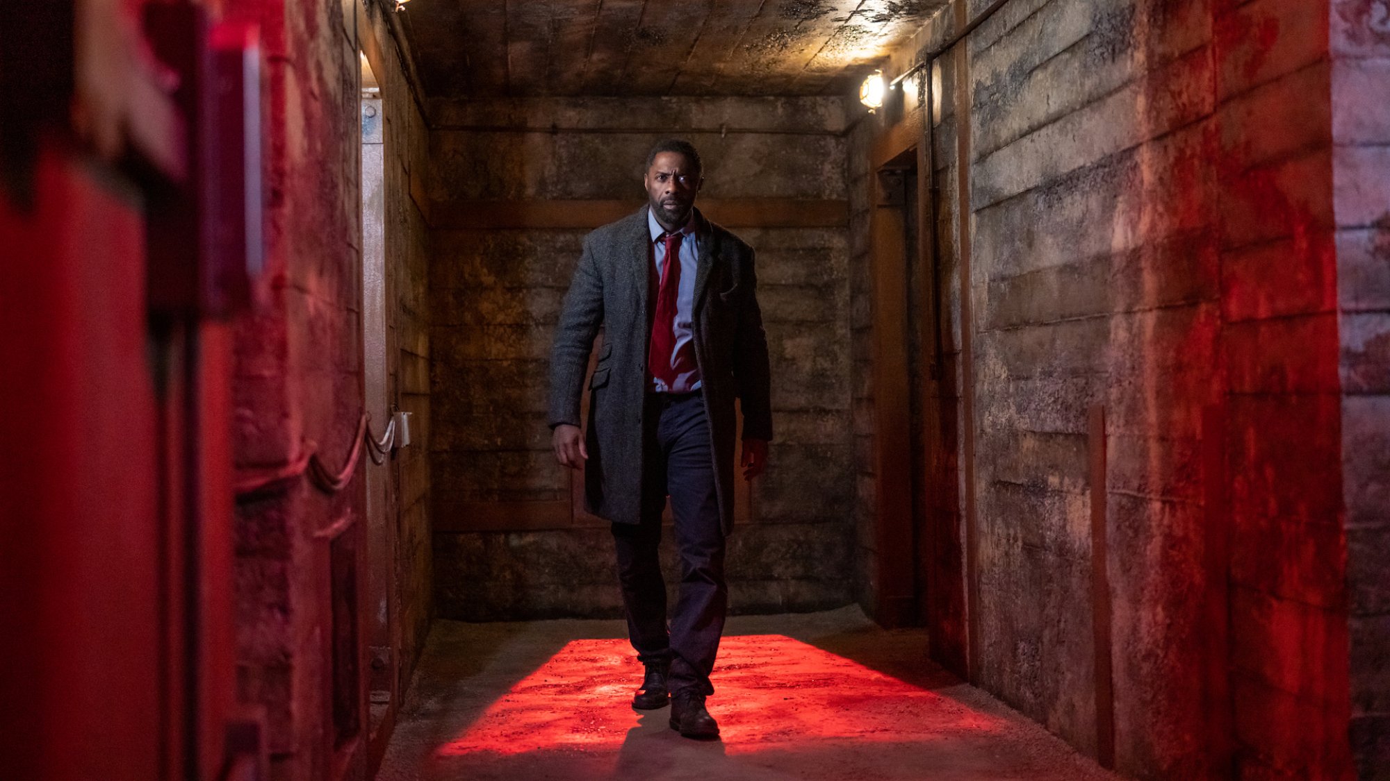 A man in a suit walks down a red-lit corridor of a shabby looking building.
