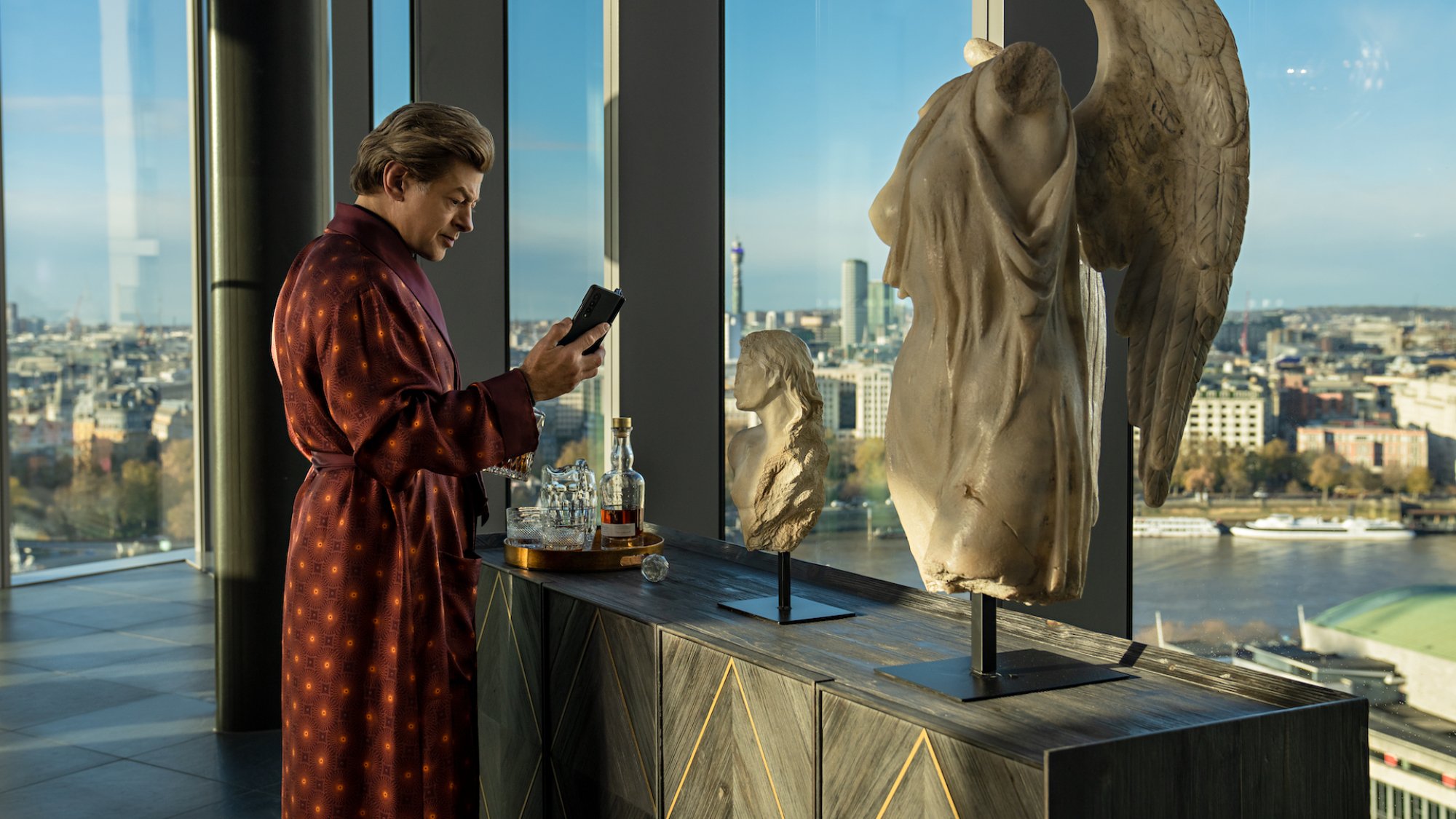 A wealthy man in a robe uses a foldable phone in an all glass apartment standing near a marble bust.