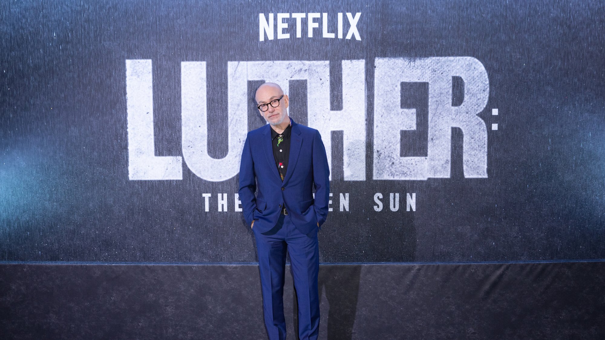 A man in a blue tailored suit and thick-rimmed glasses stands at a film premiere with the words "Netflix, Luther: The Fallen Sun" behind him.