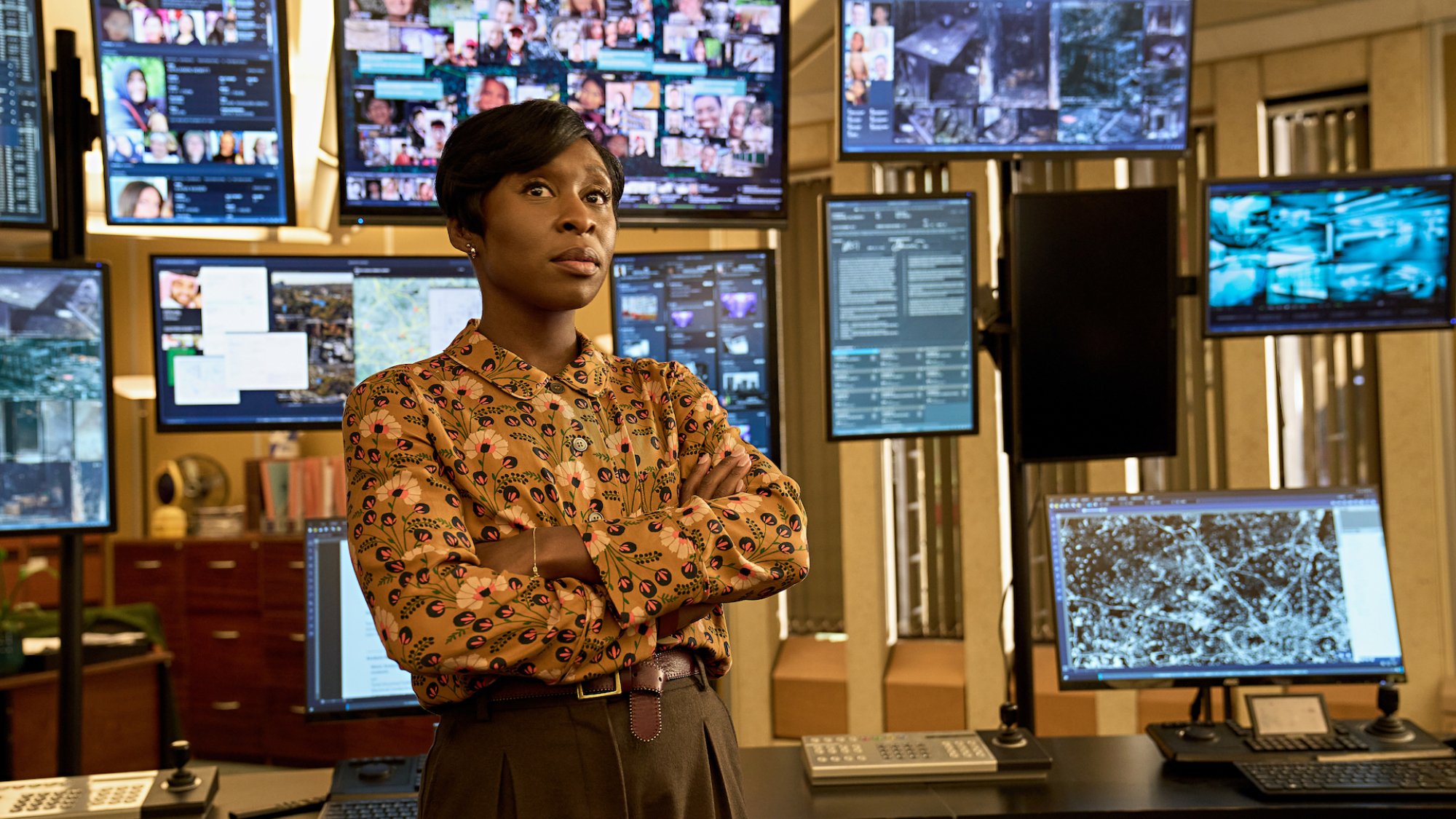 A woman in a patterned shirt stands in a police monitoring room with about 10 screens behind her showing various images and tracking information.