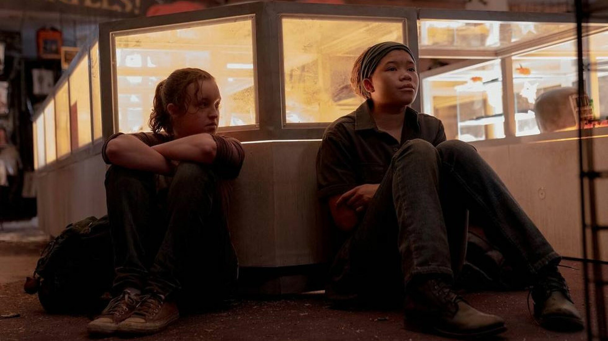 Two young girls sit down next to each other with their backs against a wall.