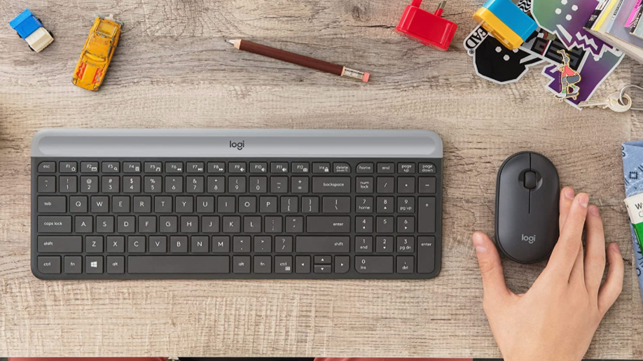 Black logitech keyboard, black logitech mouse on brown wooden desk with office supplies on the top
