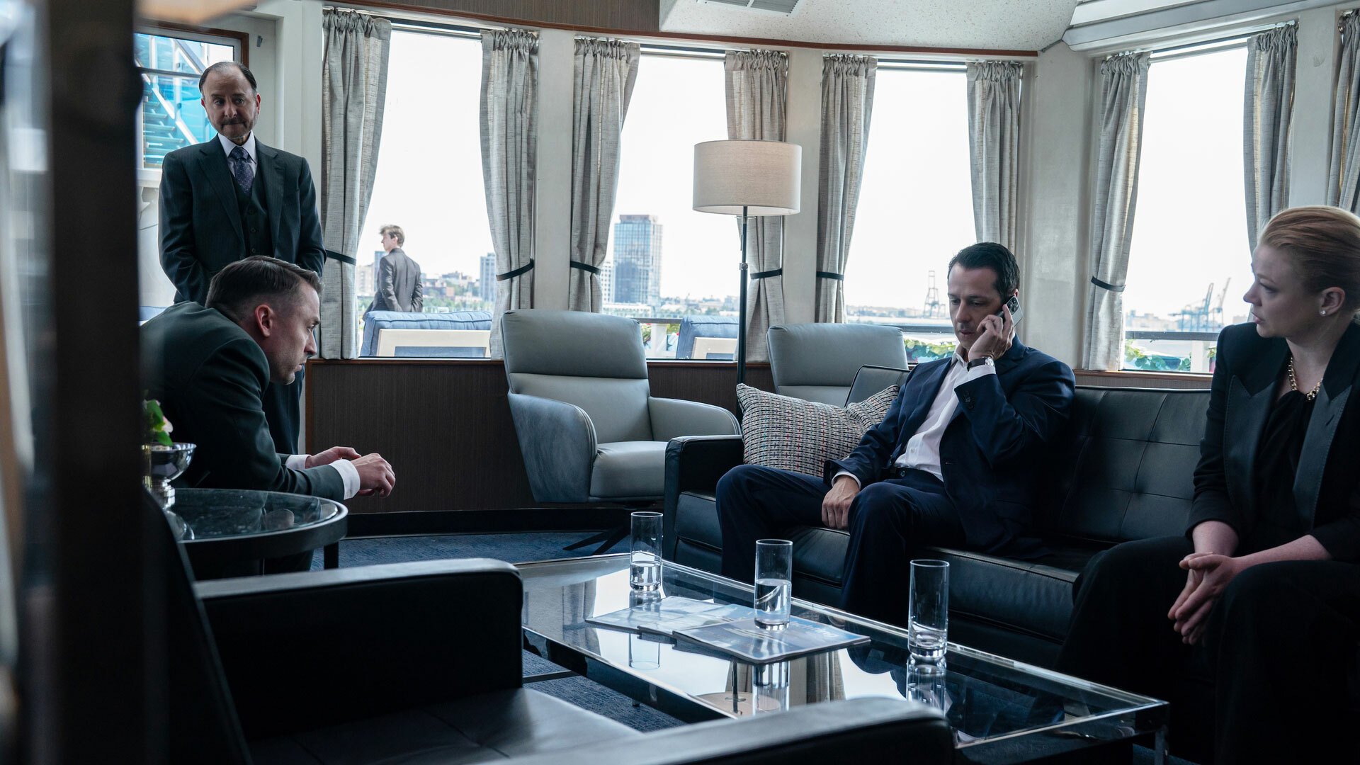 A group of people in suits sit around in a room on a boat looking sombre.