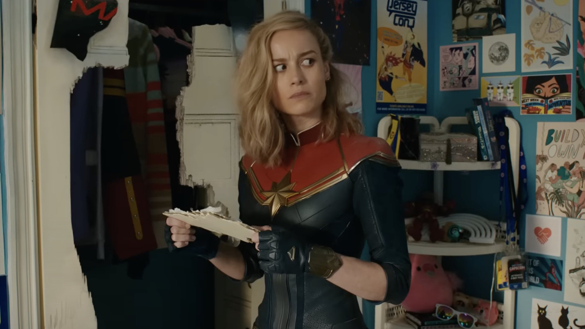 Captain Marvel stands in a teen bedroom covered in fandom for Captain Marvel.