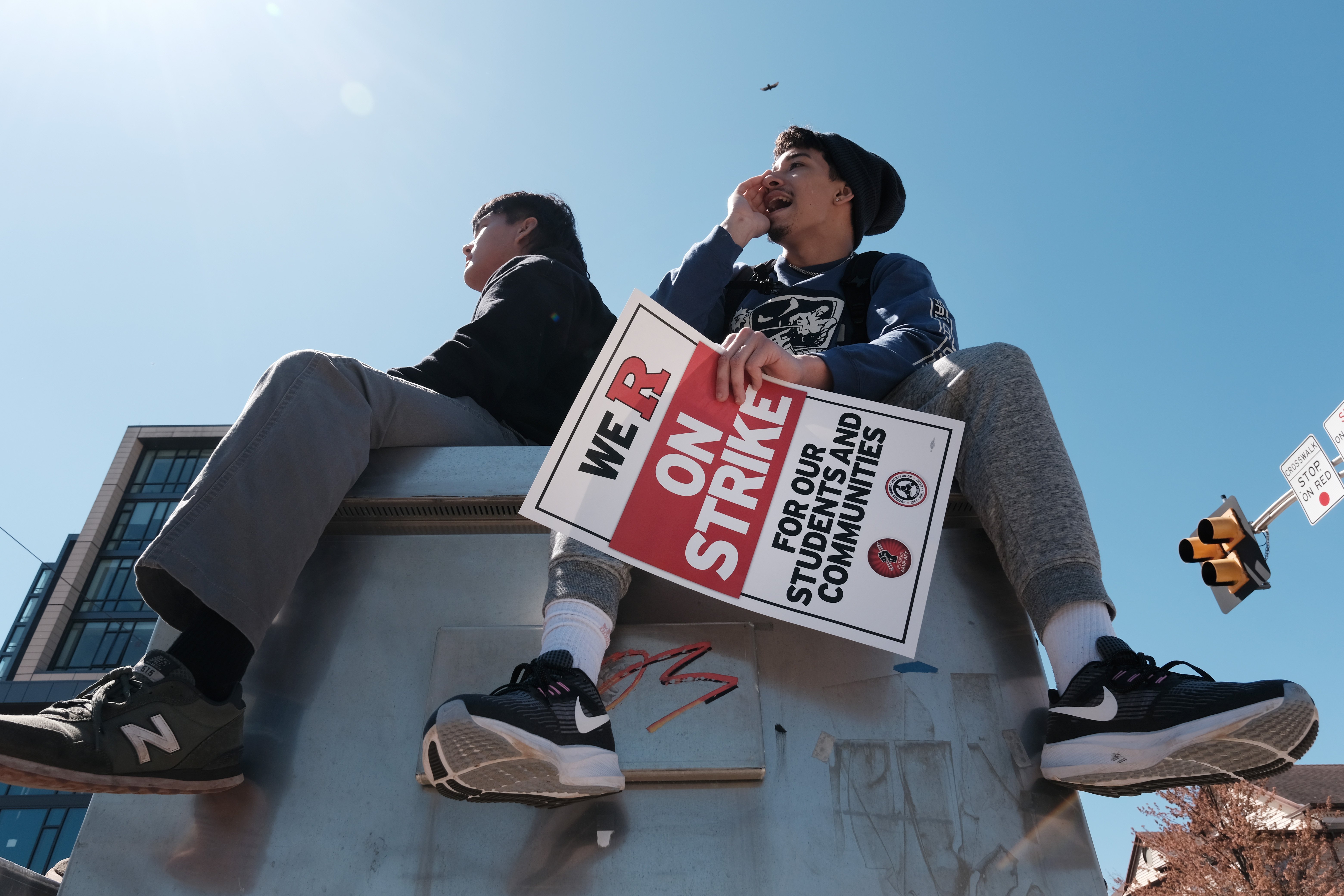 Two young people sit on top of a steel box. The one on the right dangles a red and white signs that reads,