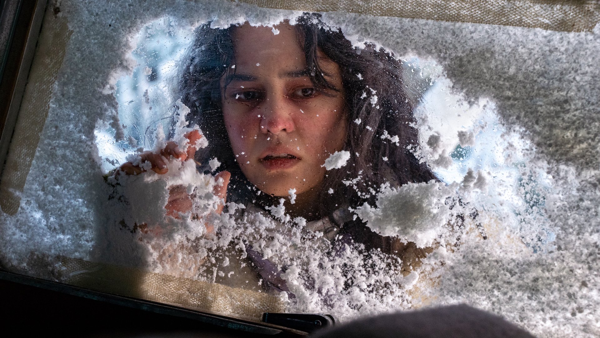 A teen girl peers through a snowy window looking concerned.