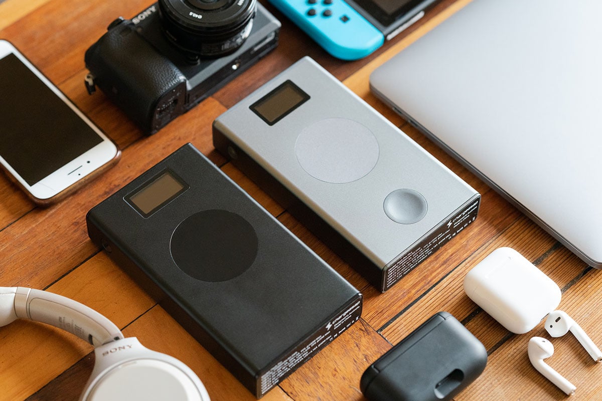 an array of devices are laid out on a wooden coffee table; the two power banks are in the center, surrounded by a camera, white headphones, an iphone, a silver laptop, and an airpods case