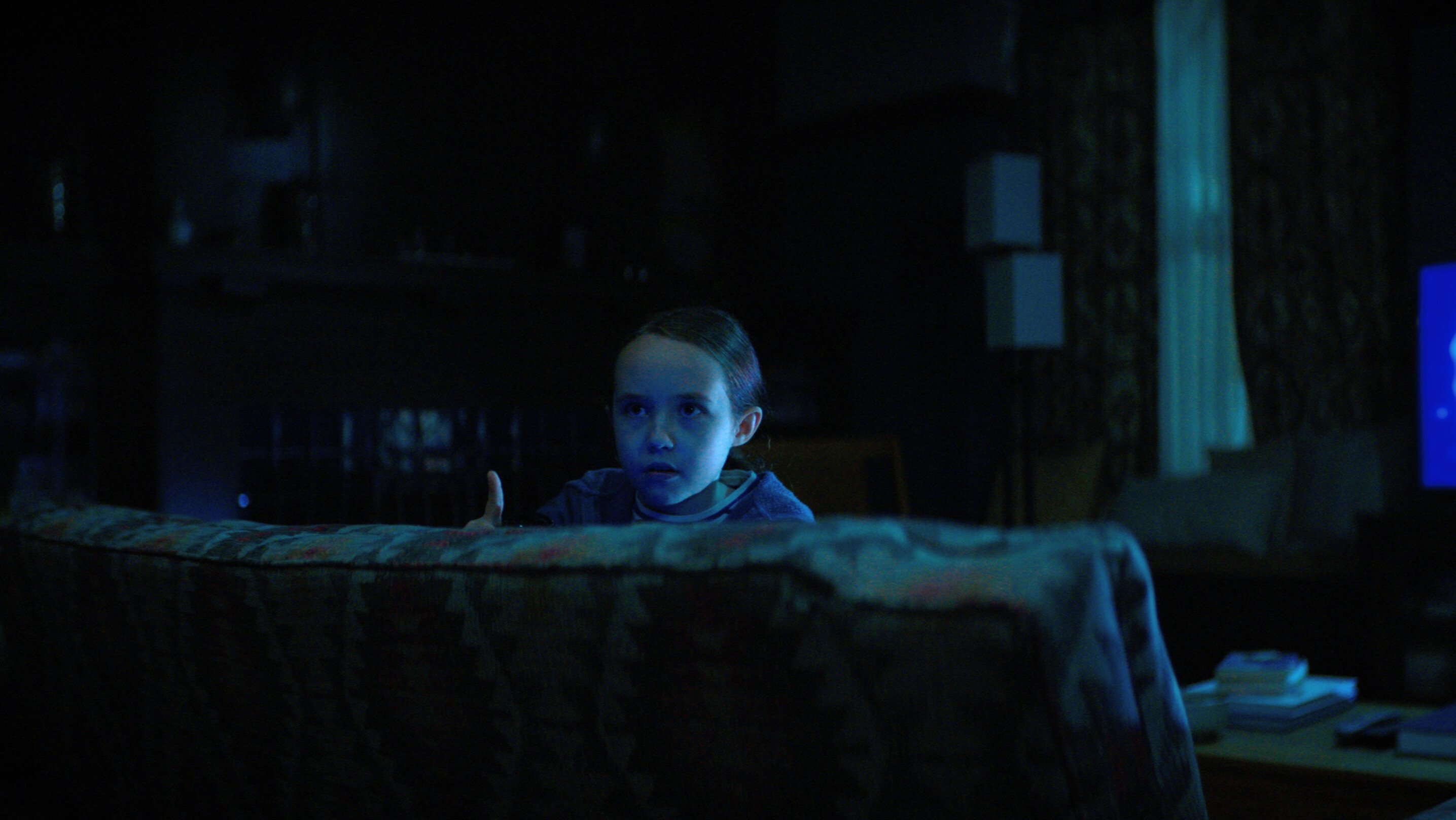 A young girl looks over a couch in a blue-lit living room.