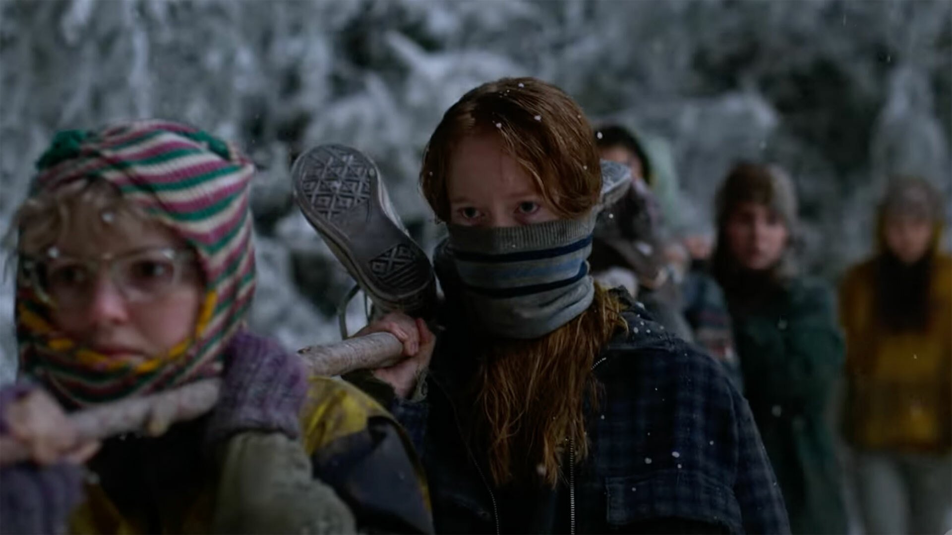 A group of teenage girls carry a body tied to a wooden pole through the snowy wilderness.