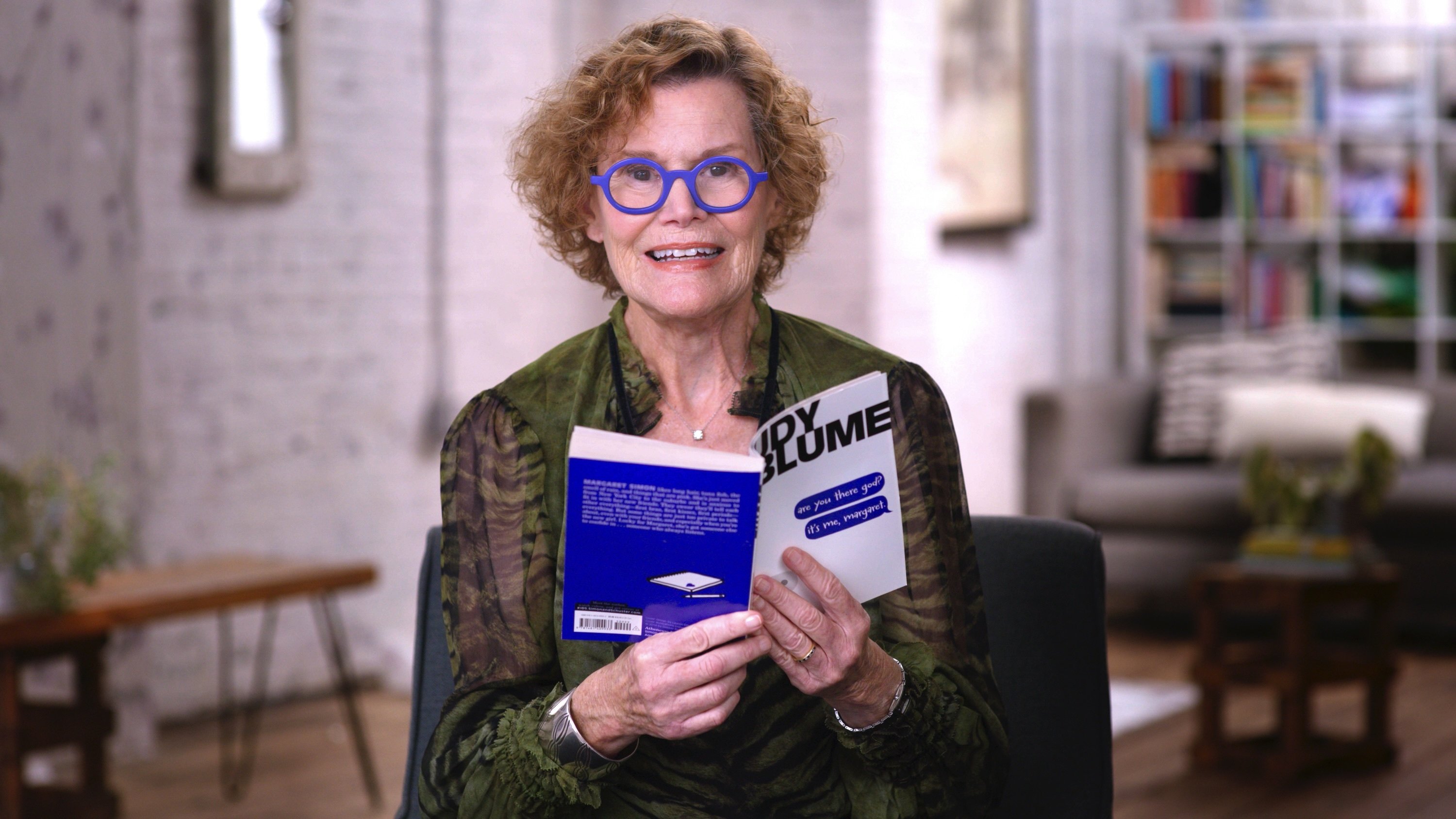 Judy Blume, wearing blue-rimmed glasses and a green blouse, reads from her book