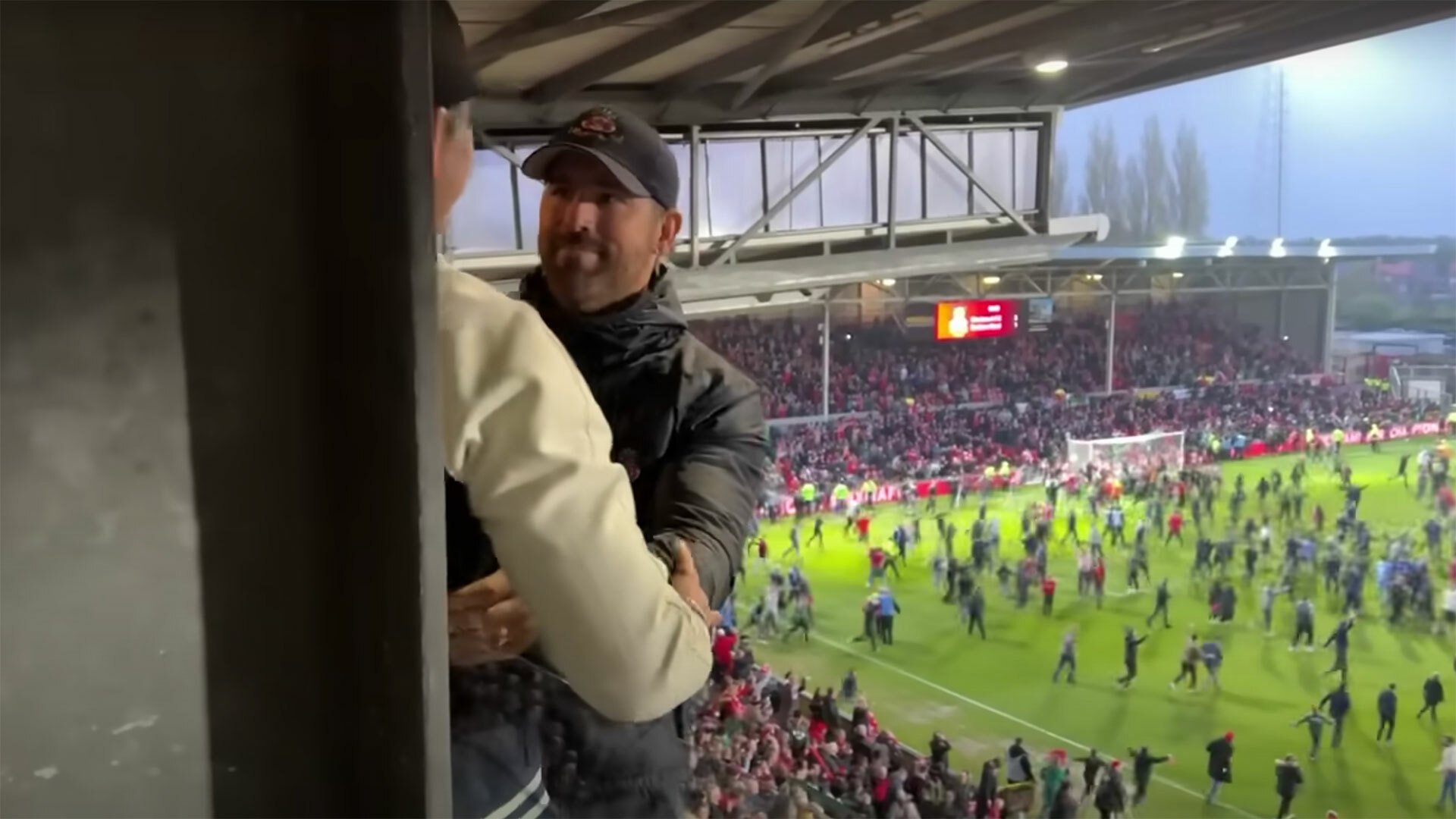 A man an the stands at a football game smiles at another man while people run onto the pitch in the background.