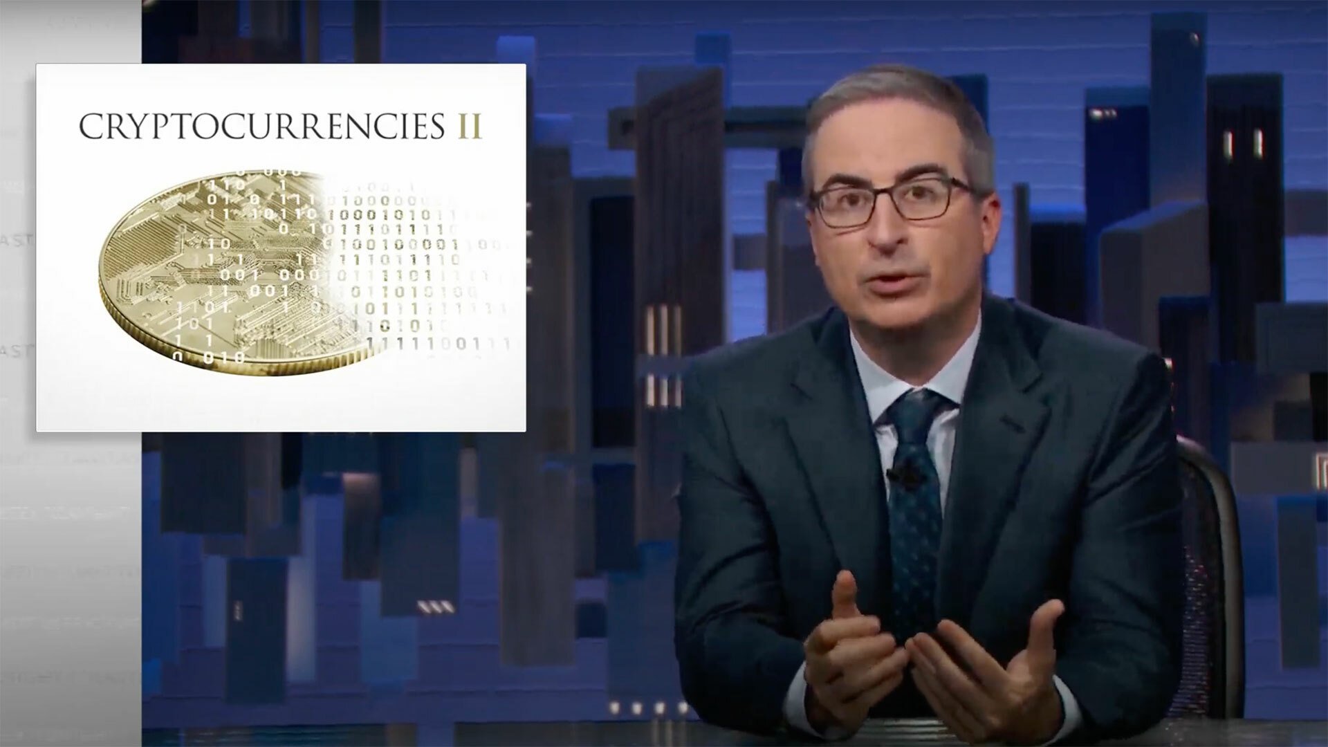 A man in glasses and a suit sits behind a desk, looking at the camera, next to an image of a coin that read 