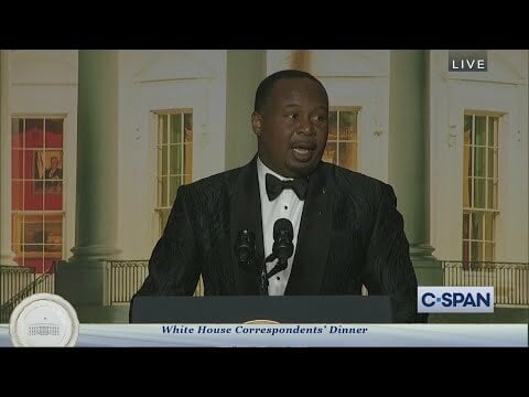 Roy Wood Jr. in a black tuxedo speaks at a podium with an image of the White House projected behind him.