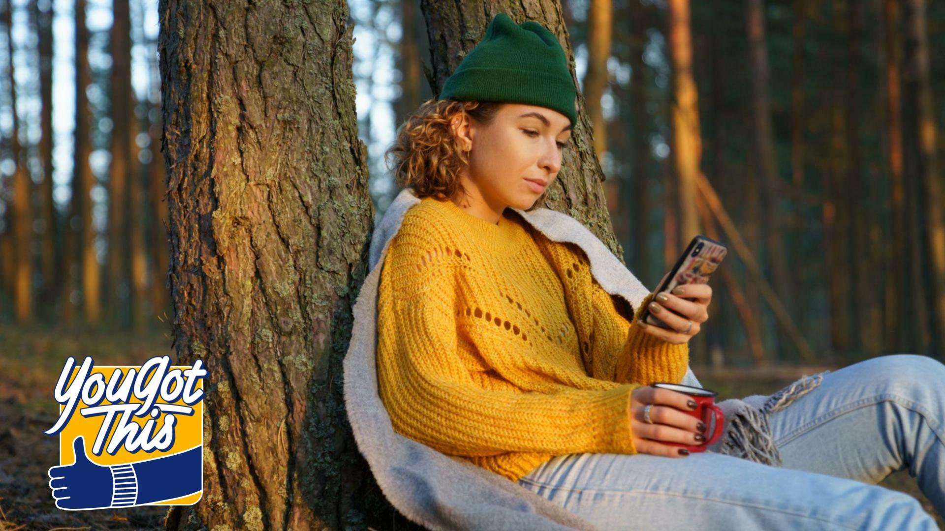 Girl sits leaning against tree with mug and looks at phone