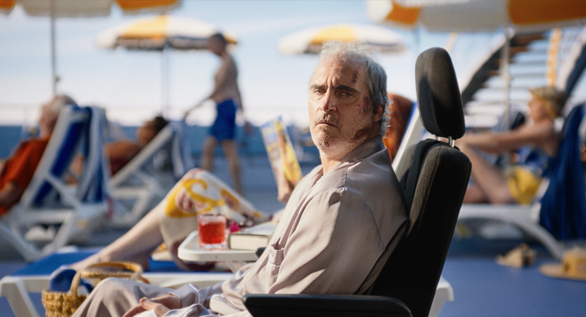 A battered-looking white man (Joaquin Phoenix) sits in an airline seat amidst people in pool chairs.