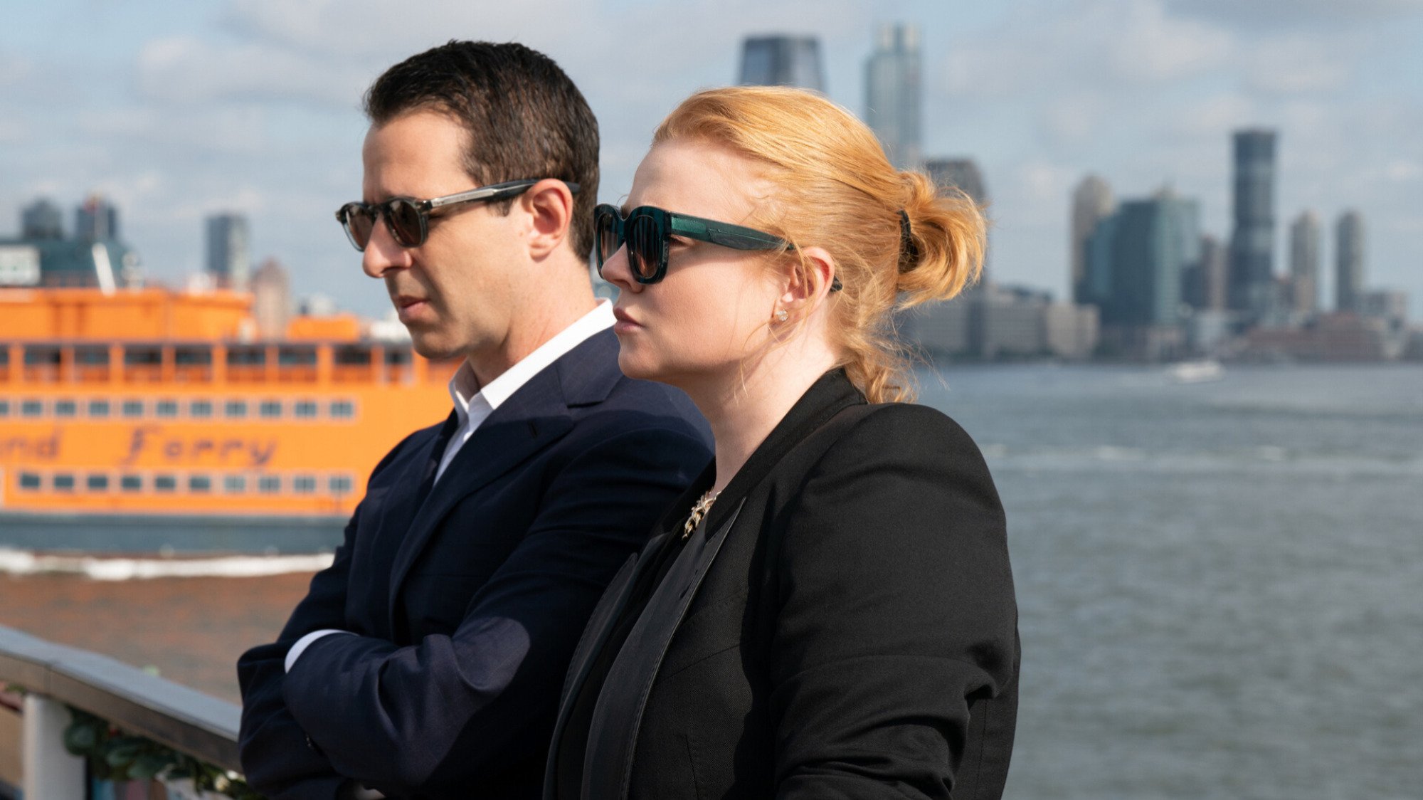 A man and a woman dressed in black suits and sunglasses stand by a boat railing.