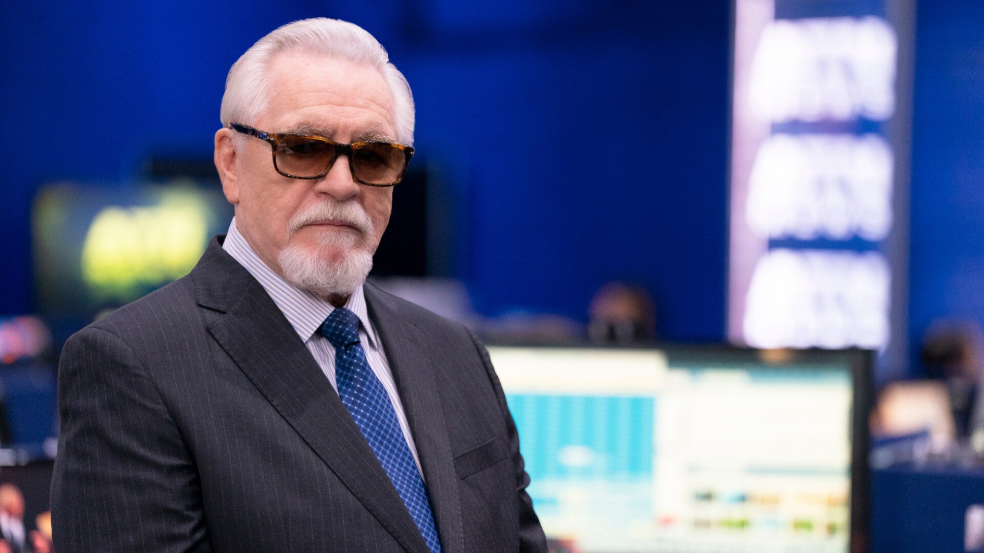 An old man wearing a suit with a blue tie stands in a news room.