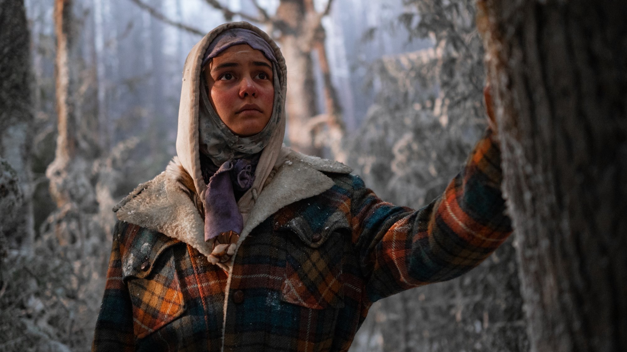 A teen girl bundled up in the snow holds her hand on a tree.
