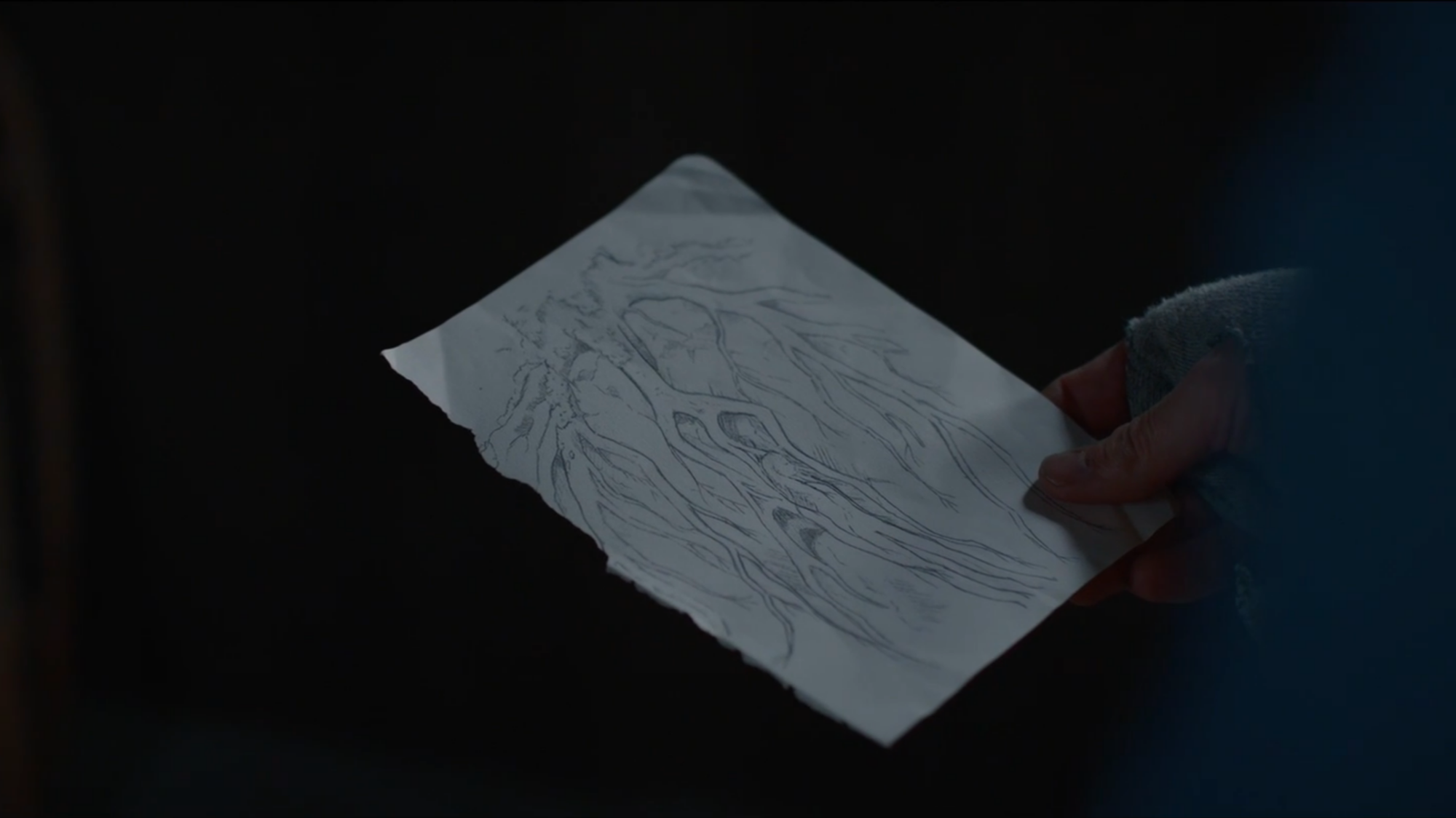 A man holds a drawing of a woman under a tree's roots.