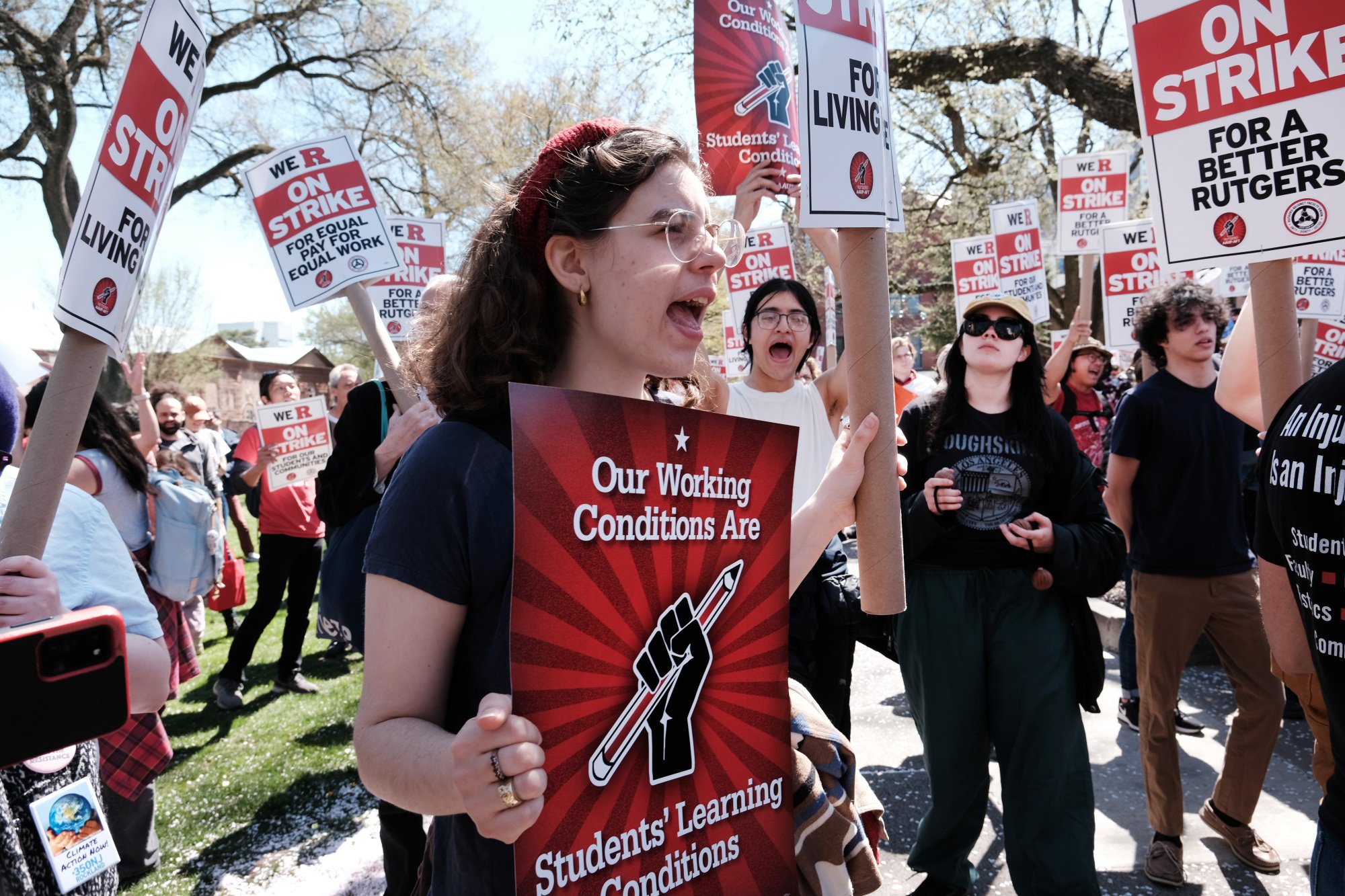 A crowd of people march with red and white "on strike" picket signs. In the foreground a person yells and holds a red sign that reads "Our working conditions are students' learning conditions".
