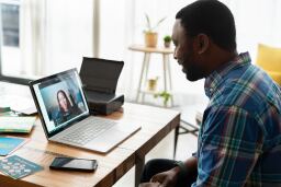man is seated at the corner of a table and taking a virtual learning course with a video instructor on his laptop
