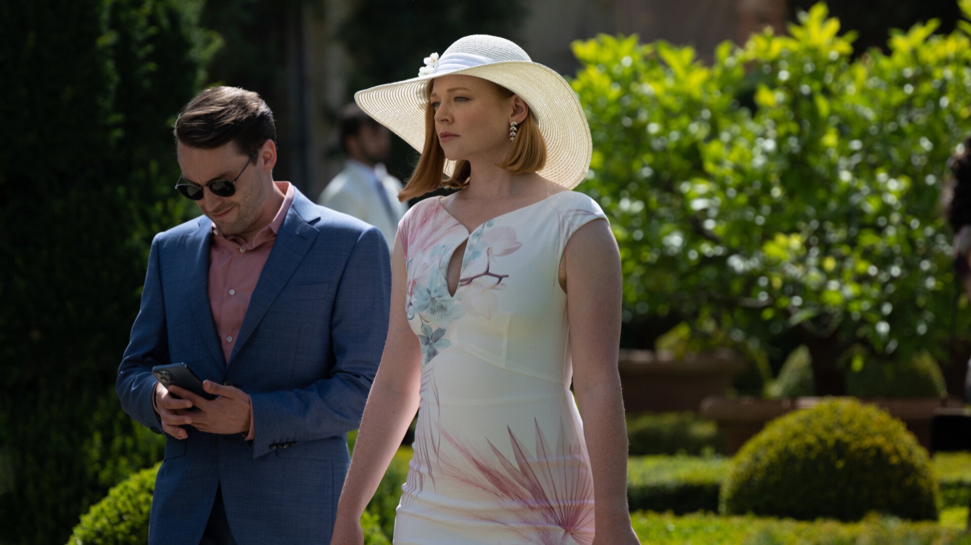 A man wearing a blue suit and a woman wearing a white floral dress walk in a garden.