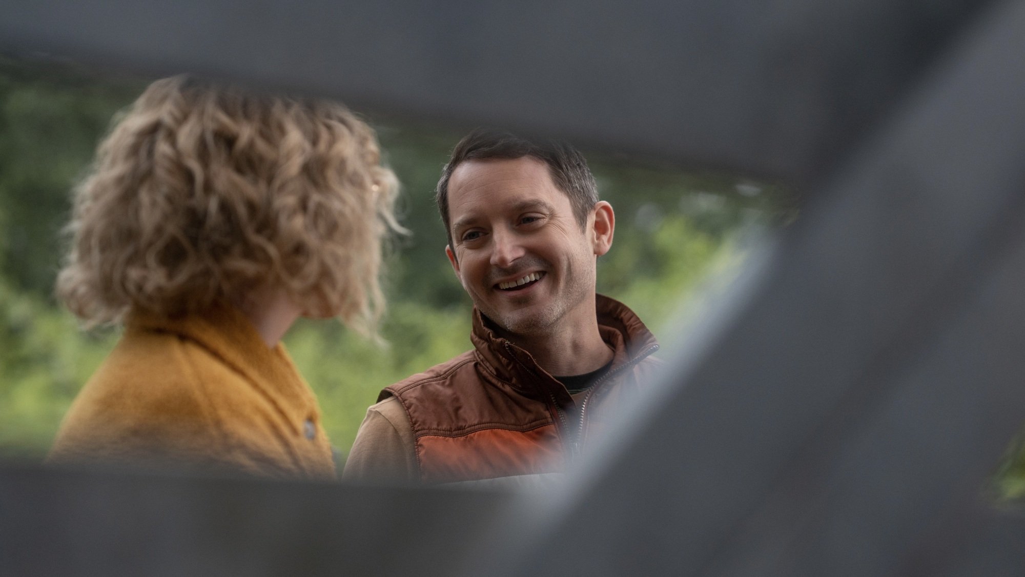 A woman in a yellow coat and a smiling man in a brown and orange vest peer through the slats of a fence.
