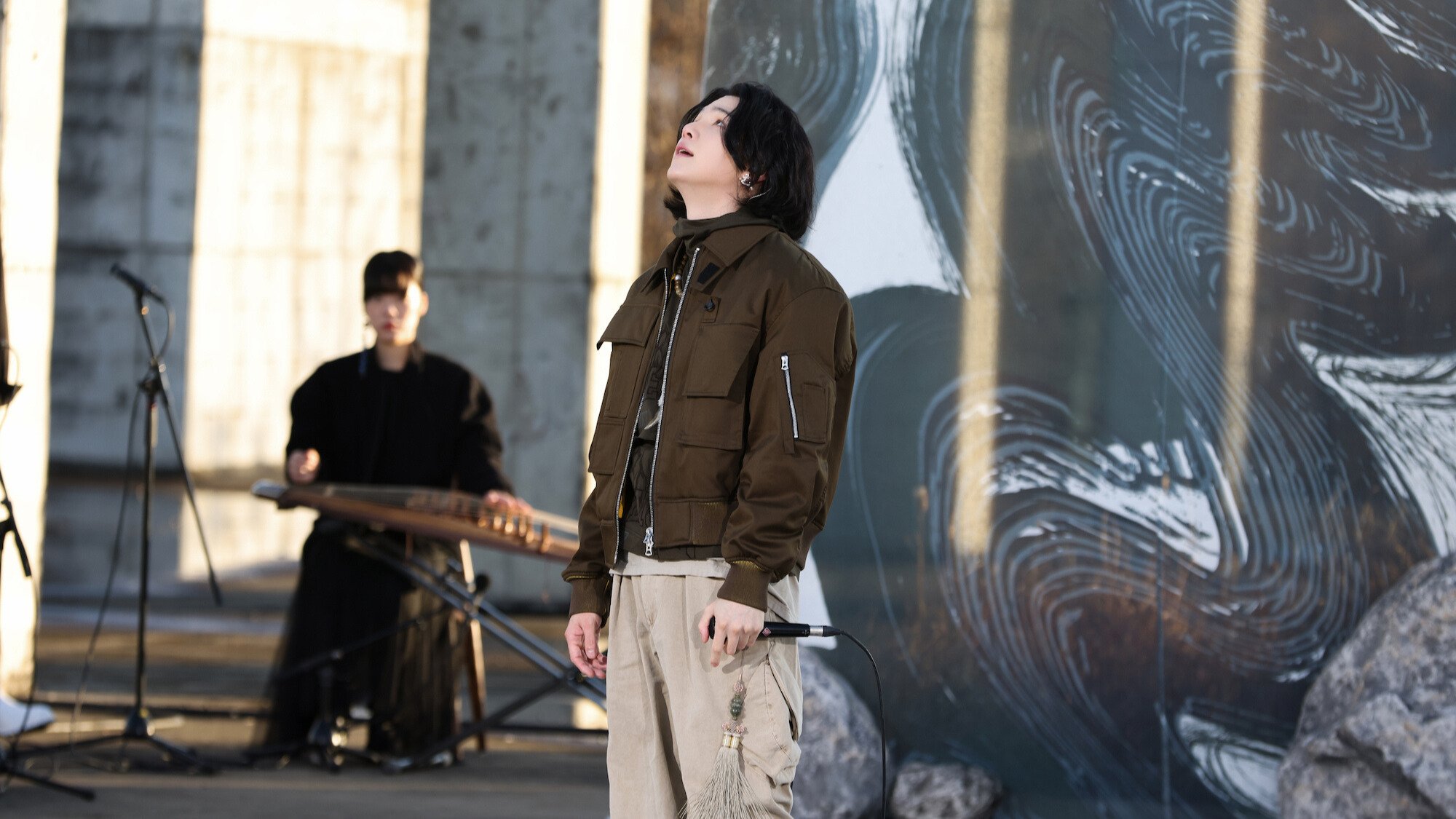 Suga from BTS performs outdoors in front of a black mural painting.