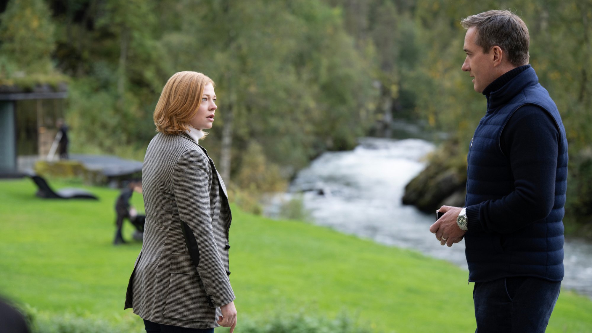 In "Succession" a woman and a man in smart clothes stand in tense conversation by a river.