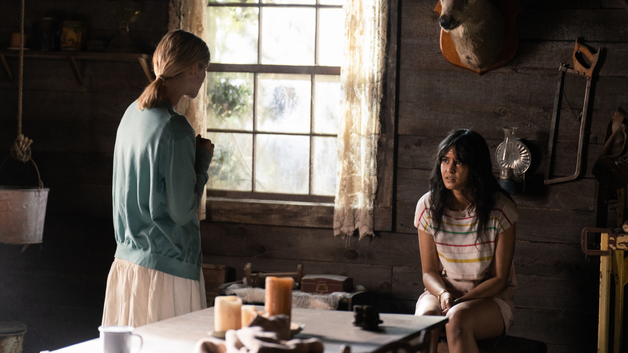 Two teen girls, one sitting, one standing, talk in a cabin kitchen.