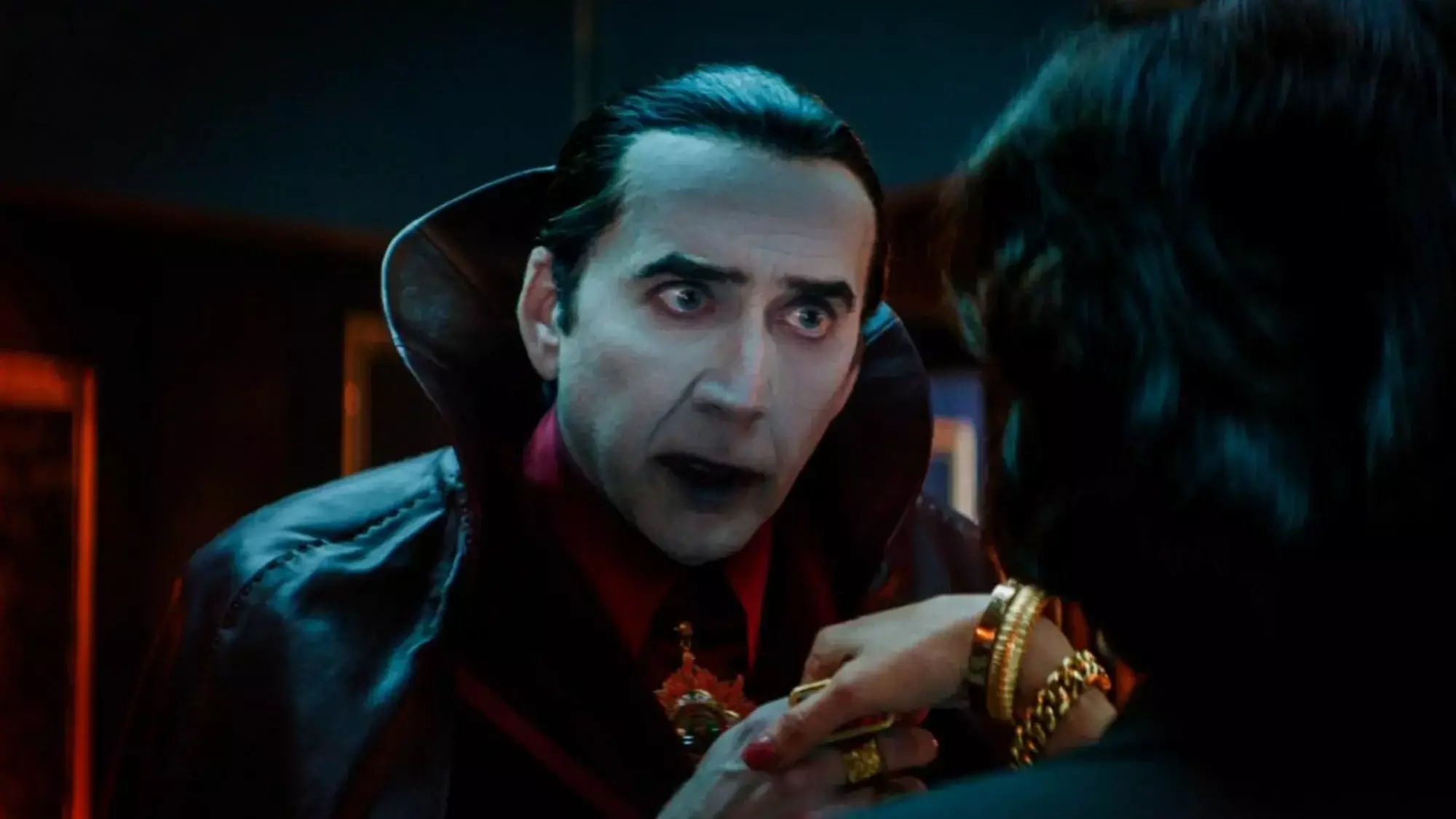 Nicolas Cage plays Dracula in "Renfield," with slicked-back hair, a white-painted face, and red lips, clutching a woman's hand.