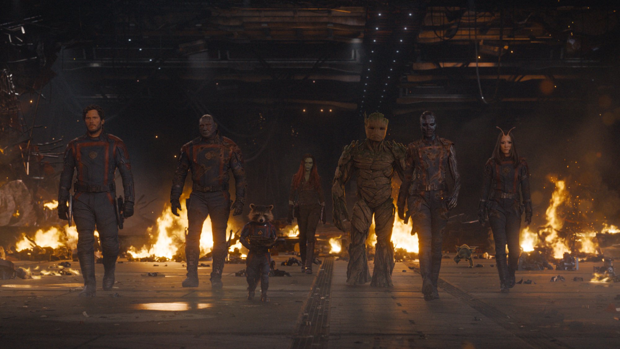 The Guardians all march in a line away from a burning structure.